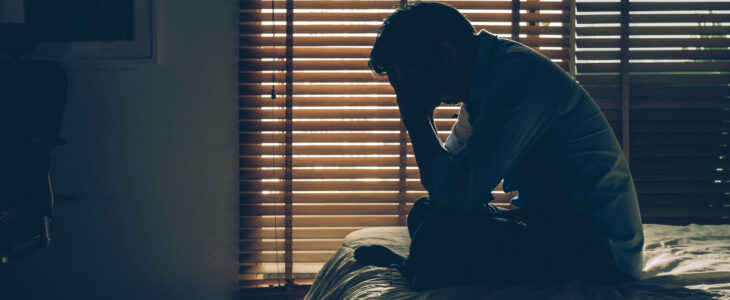Sad man sitting head in hands on the bed in the dark bedroom with low light